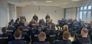 Na sali wykładowej tyłem do zdjęcia siedzą w rzędach umundurowani policjanci. Przed nimi skierowani twarzą stoją w odstępach policjanci prowadzący szkolenie.